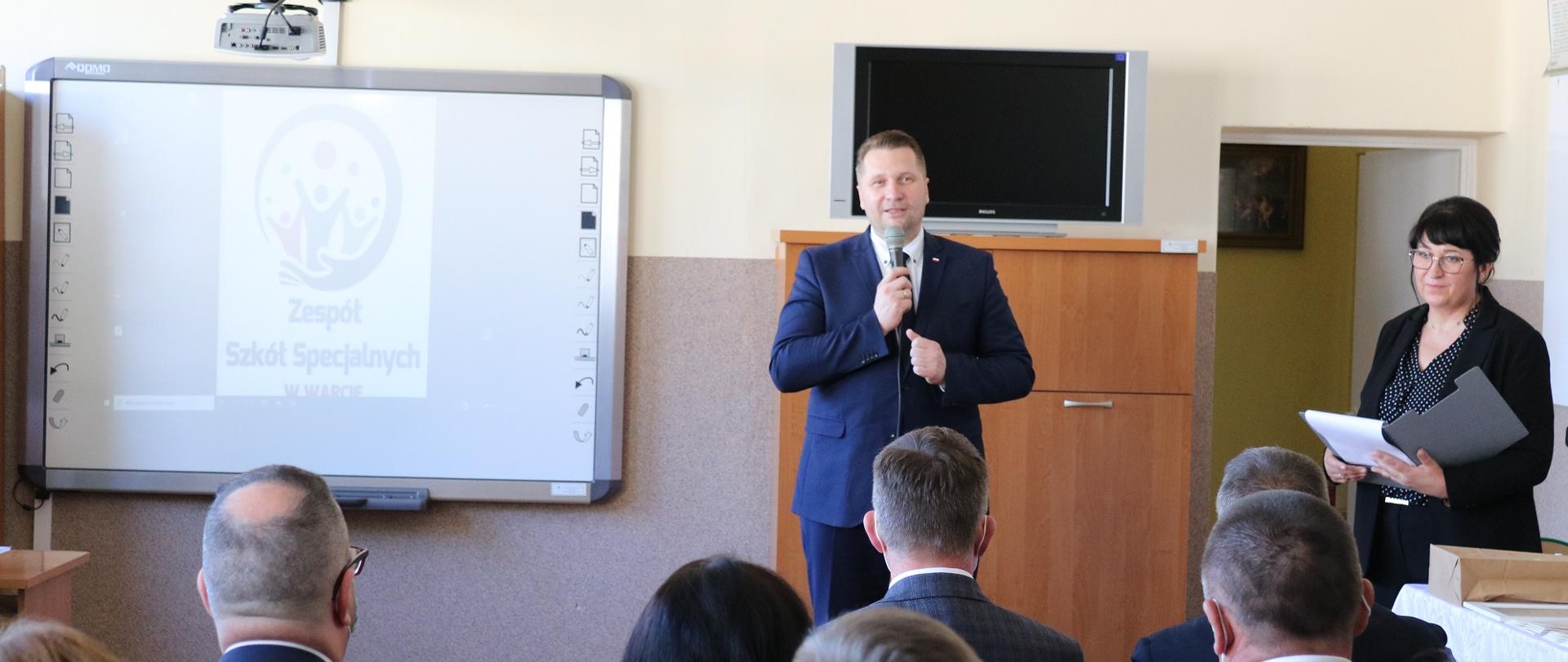 Minister w klasie szkolnej przemawia do osób siedzących w sali lekcyjnych.