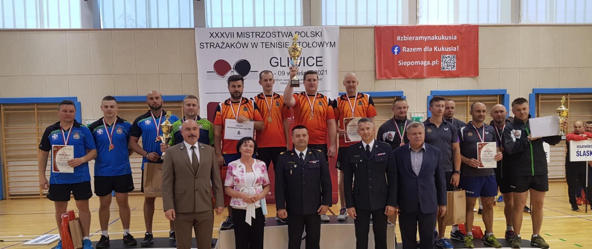 Zdjęcie grupowe, zwycięzcy na podium, przed nimi stoi 2 funkcjonariuszy PSP oraz trzy inne osoby