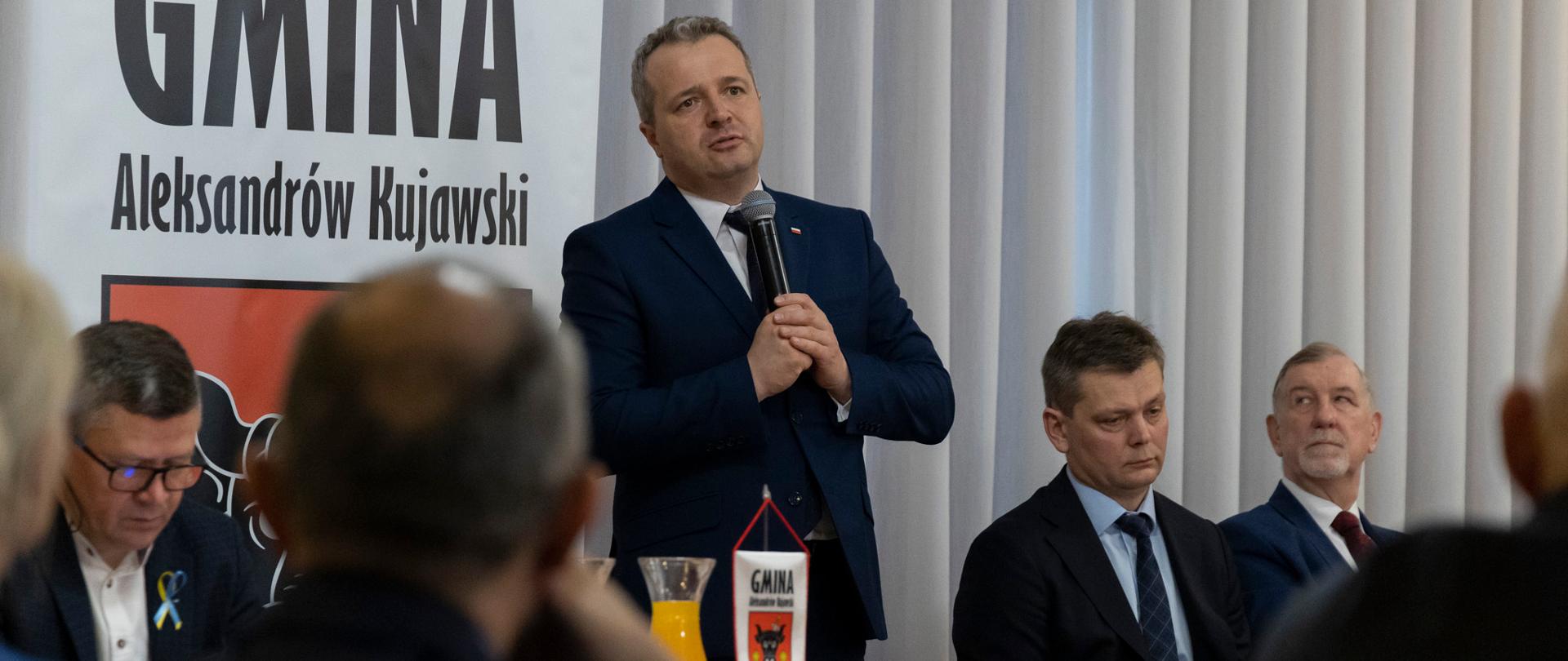 Jeden mężczyzna stojący z mikrofonem i wypowiadający się, kilka osób siedzących przy stole