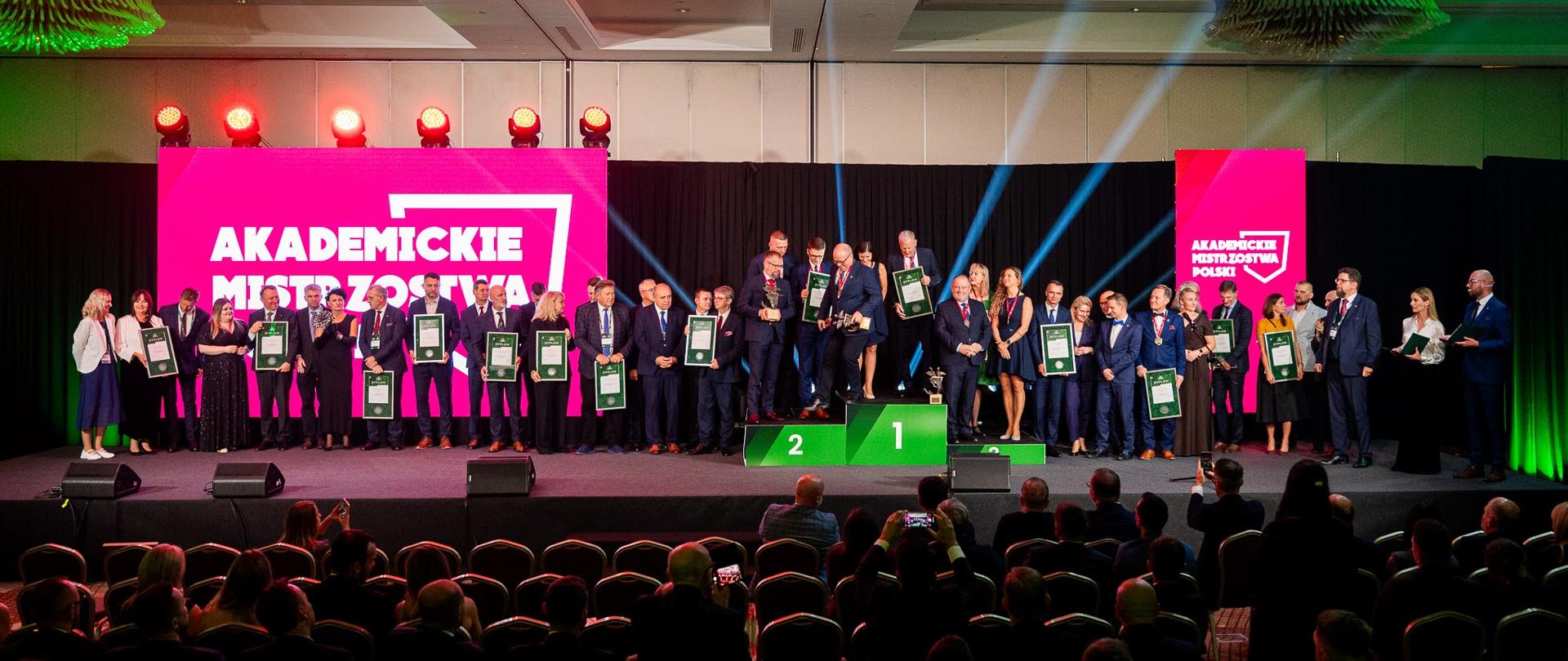 Widok na wielką salę, na podwyższeniu stoi duża grupa ludzi, przed nimi na zielonym podium trzy osoby, w tle pod ścianą zasłoniętą czarnym materiałem różowe ekrany z napisem Akademickie Mistrzostwa Polski.