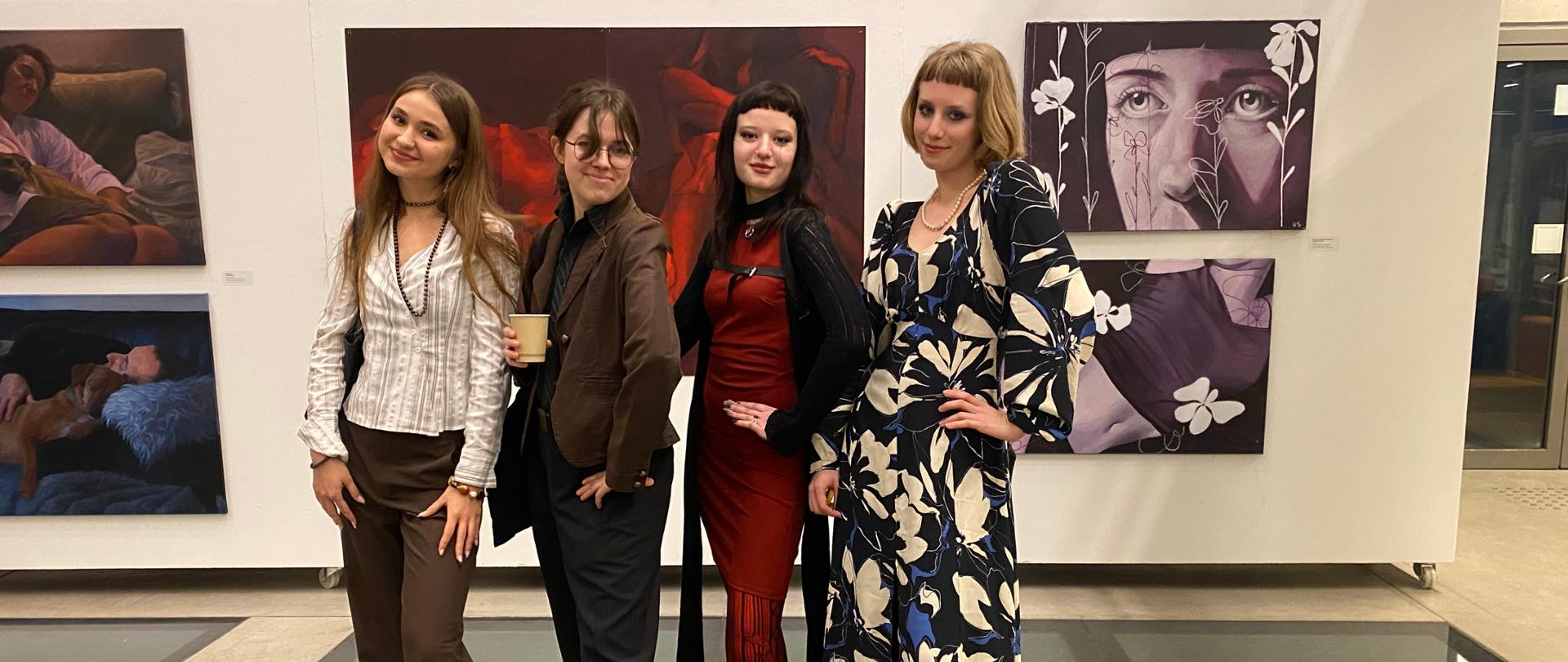 Cztery dziewczyny, stojące rzędem, pozują do zdjęcia na tle ekspozycji malarskiej