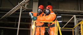 Dwóch strażaków w pomarańczowych ubraniach przeciwchemicznych, w aparatach i maskach powietrznych oraz czerwonych hełmach wykonuje uszczelnienie rozszczelnionej rury w budynku zakładu.