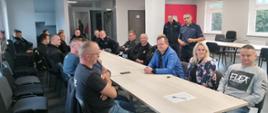 Zdjęcie przedstawia strażaków PSP i druhów OSP siedzących przy stole w sali Komendy Powiatowej Państwowej Straży Pożarnej w Człuchowie. Na środku sali stoją policjanci prowadzący szkolenie z zakresu przepisów ruchu drogowego.