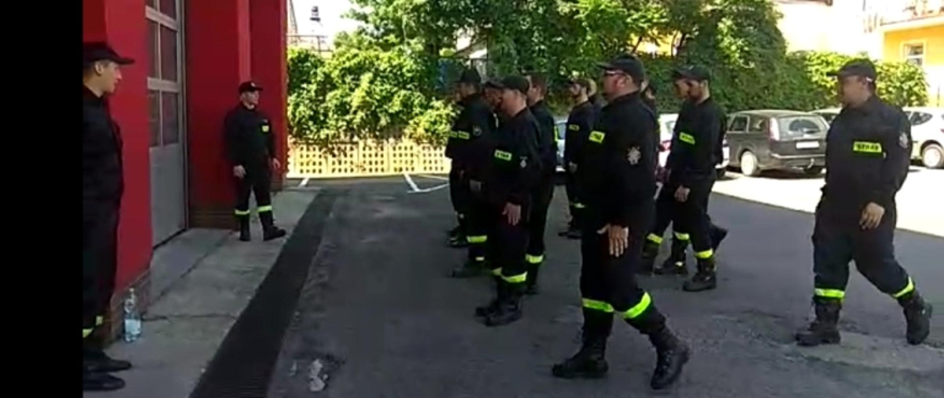 Kolorowe zdjęcie przedstawia grupę umundurowanych strażaków na placu przed budynkiem strażnicy. Strażacy podczas ćwiczeń w grupie.