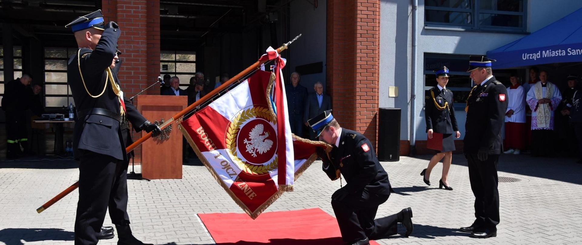 Zdjęcie wykonane na zewnątrz. Przedstawia poczet sztandarowy oraz Komendanta który ukląkł i całuje sztandar.