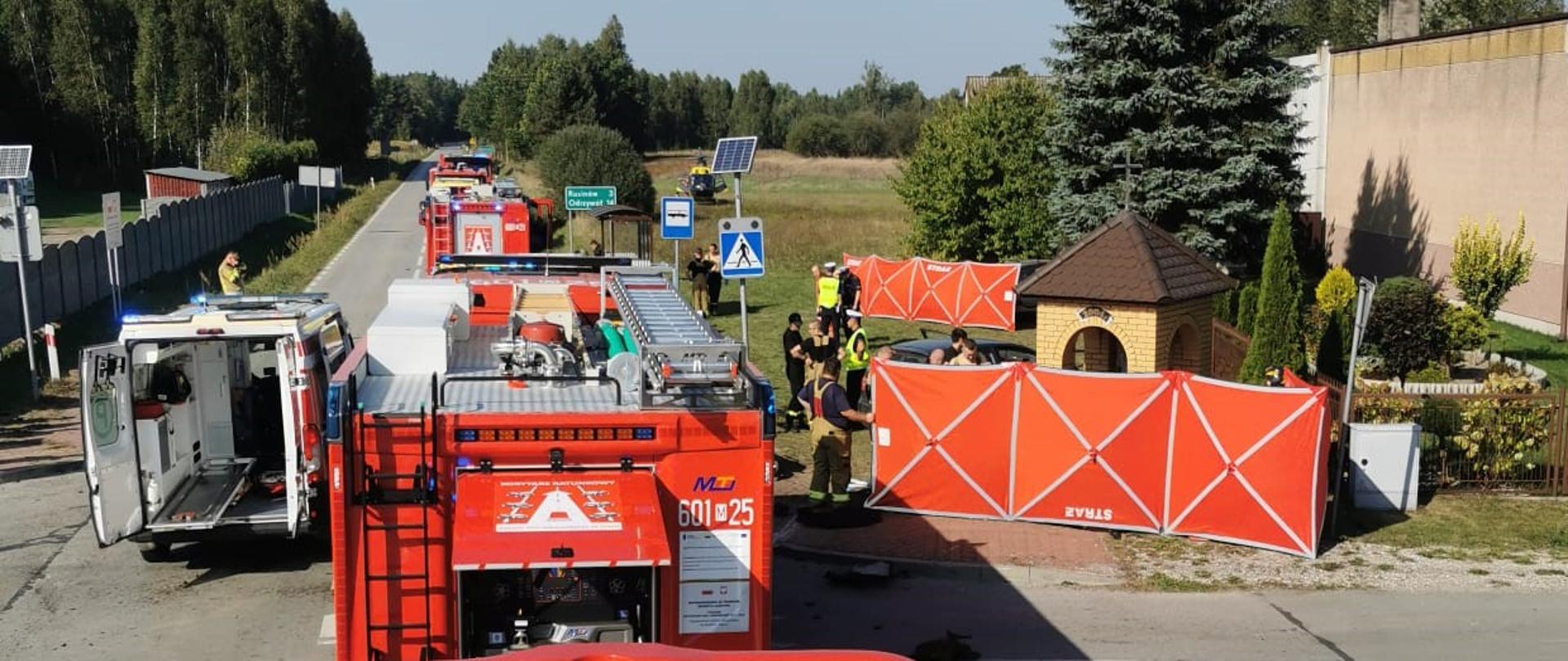 Zdjęcie przedstawia scenę wypadku lub zdarzenia drogowego, widzianą z tyłu kabiny czerwonego samochodu ciężarowego, prawdopodobnie kolejnego wozu strażackiego, na którym widoczny jest numer "609M69". Akcja rozgrywa się w słoneczny dzień, na drodze biegnącej przez obszar częściowo zalesiony lub wiejski. W centralnej części kadru, na drodze, stoją dwa czerwone wozy strażackie z numerami "601 25" i "609M69". Jeden z nich ma otwarte boczne drzwi, ukazujące wnętrze. Wokół pojazdów i na poboczu zgromadzeni są strażacy w żółtych kombinezonach i hełmach, a także inni ludzie. Widoczne są również elementy zabezpieczające teren, w tym biało-niebieska chorągiewka ostrzegawcza i czerwono-biała płachta, prawdopodobnie parawan, częściowo zasłaniająca obszar zdarzenia. W tle widać wiejskie zabudowania, drzewa iglaste i liściaste oraz jasne niebo. Cała scena wskazuje na trwającą interwencję służb ratunkowych po wypadku drogowym.