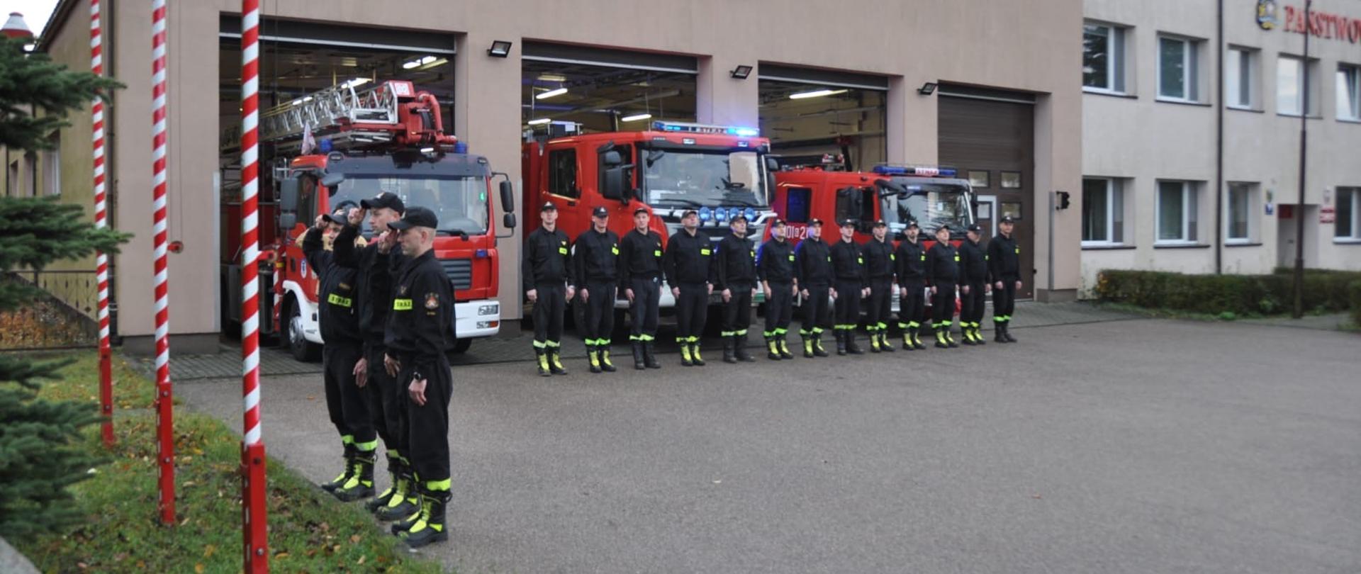Strażacy ubrani w czarne ubrania koszarowe, stoją na tle czerwonych pojazdów pożarniczych. 
