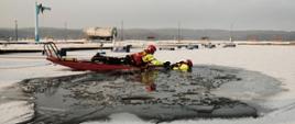 Słoneczny dzień. Teren mariny w Charzykowach. Na zamarzniętym jeziorze strażak zabezpieczony liną asekuracyjną na saniach lodowych wyciąga z wody drugiego strażaka, który znajduje się w wodzie w przeręblu. W oddali widać pomosty oraz las po drugiej stronie jeziora.