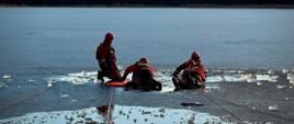 Na zamarzniętym jeziorze prowadzone są ćwiczenia ratownictwa lodowego. Na pierwszym planie widać rozkruszony lód i przerębel z ciemną wodą. Dwóch ratowników w czerwonych, specjalistycznych ubraniach wypornościowych leży na czerwonej desce lodowej. Ratownicy są w kaskach i kamizelkach asekuracyjnych. Z prawej strony trzeci ratownik w ubraniu strażackim ciągnie deskę za pomocą linki ratowniczej. W tle widoczny jest pomost oraz zamarznięta tafla jeziora i drzewa.