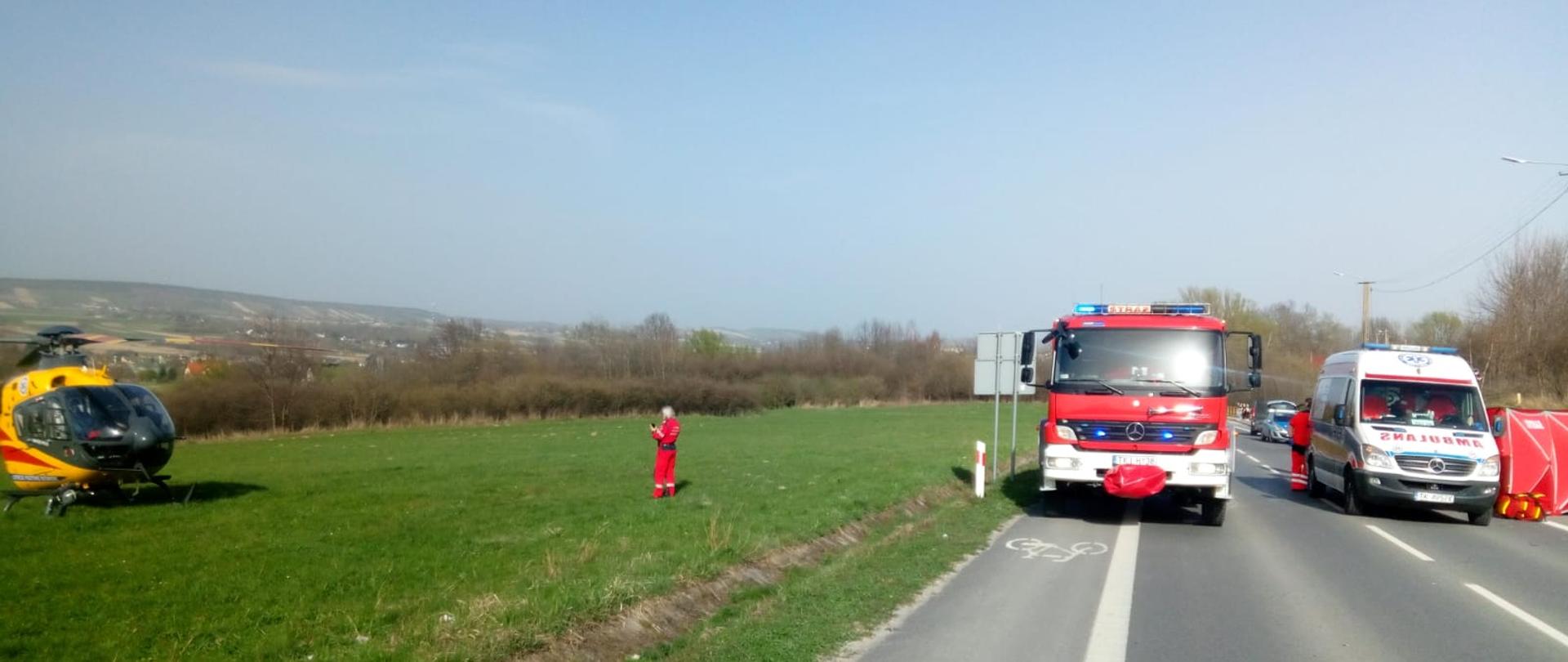 Zdjęcie przedstawia stojące na drodze wojewódzkiej samochód pożarniczy i po prawej ambulans. Za ambulansem widać rozłożony czerwony parawan osłaniający, a po lewej stronie, na pobliskiej łące wylądował helikopter do którego zbliża się ratownik.