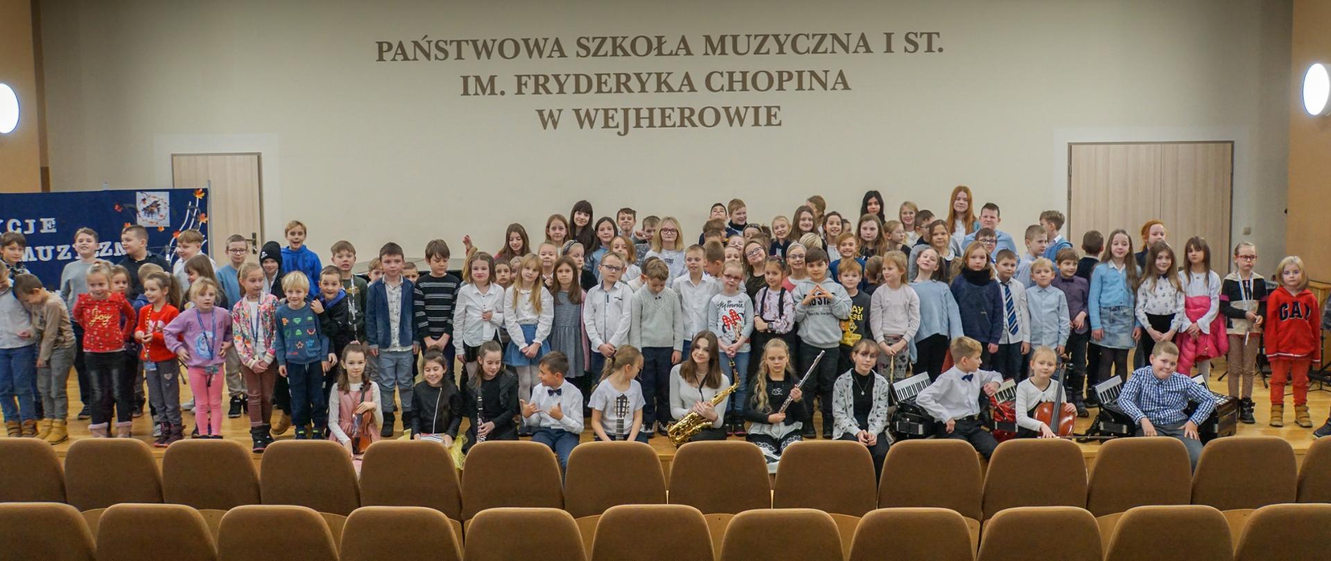 Zdjęcie grupowe na którym około setka dzieci ze szkoły podstawowej pozuje na estradzie szkolnej sali koncertowej po audycji umuzykalniającej. Na pierwszym planie siedzą wykonawcy koncertu w ilości 11 osób.