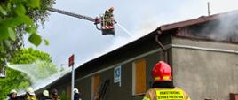 Na palący się budynek podawany jest prąd gaśniczy z drabiny oraz podnośnika, wokół budynku widać pracujących strażaków