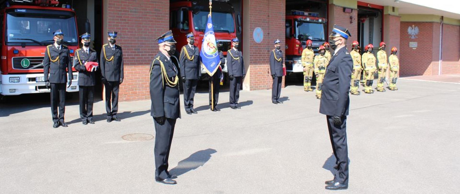 Na tle budynku Komendy Miejskiej PSP w Łomży widoczni są funkcjonariusze PSP podczas uroczystego apelu stojący na baczność. Dowódca uroczystości składa meldunek Komendantowi Miejskiemu PSP w Łomży. Strażacy ubrani są w mundury wyjściowe oraz ubranie specjalne. 