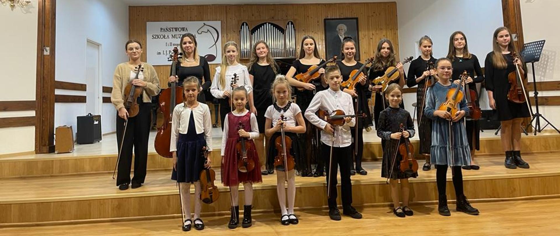 Fotografia przedstawiająca uczestników wydarzenia ubranych na galowo stojących w dwóch rzędach na scenie szkoły muzy znej, trzymających instrumenty.