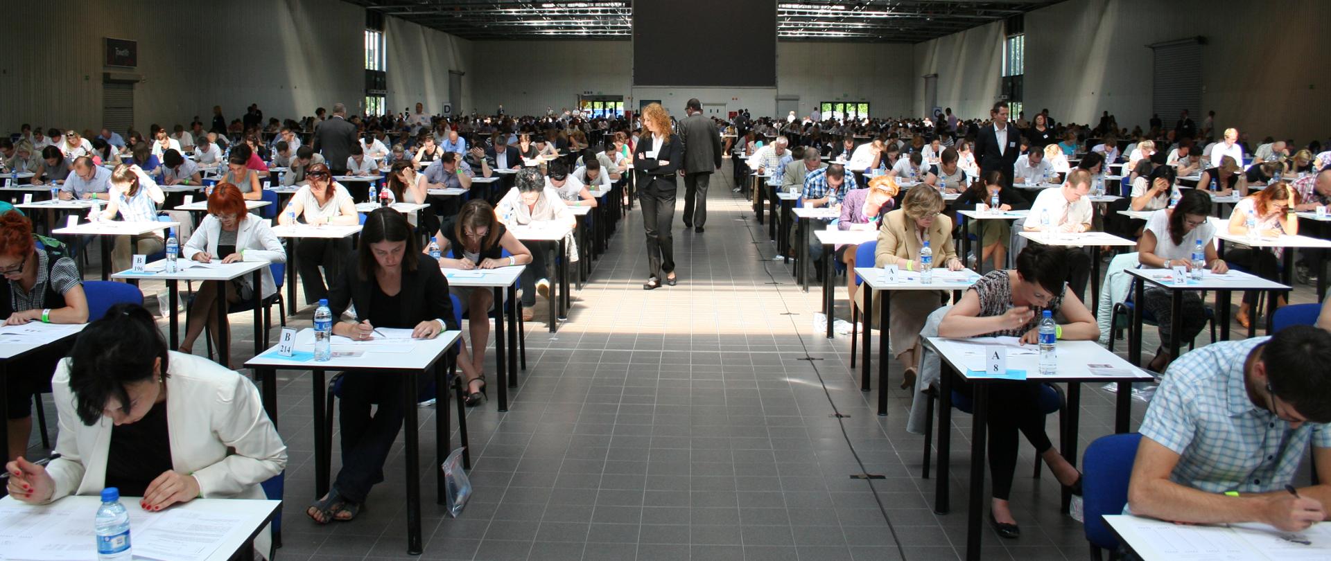 Uczestnicy postępowania kwalifikacyjnego siedzą przy stolikach w dużej hali