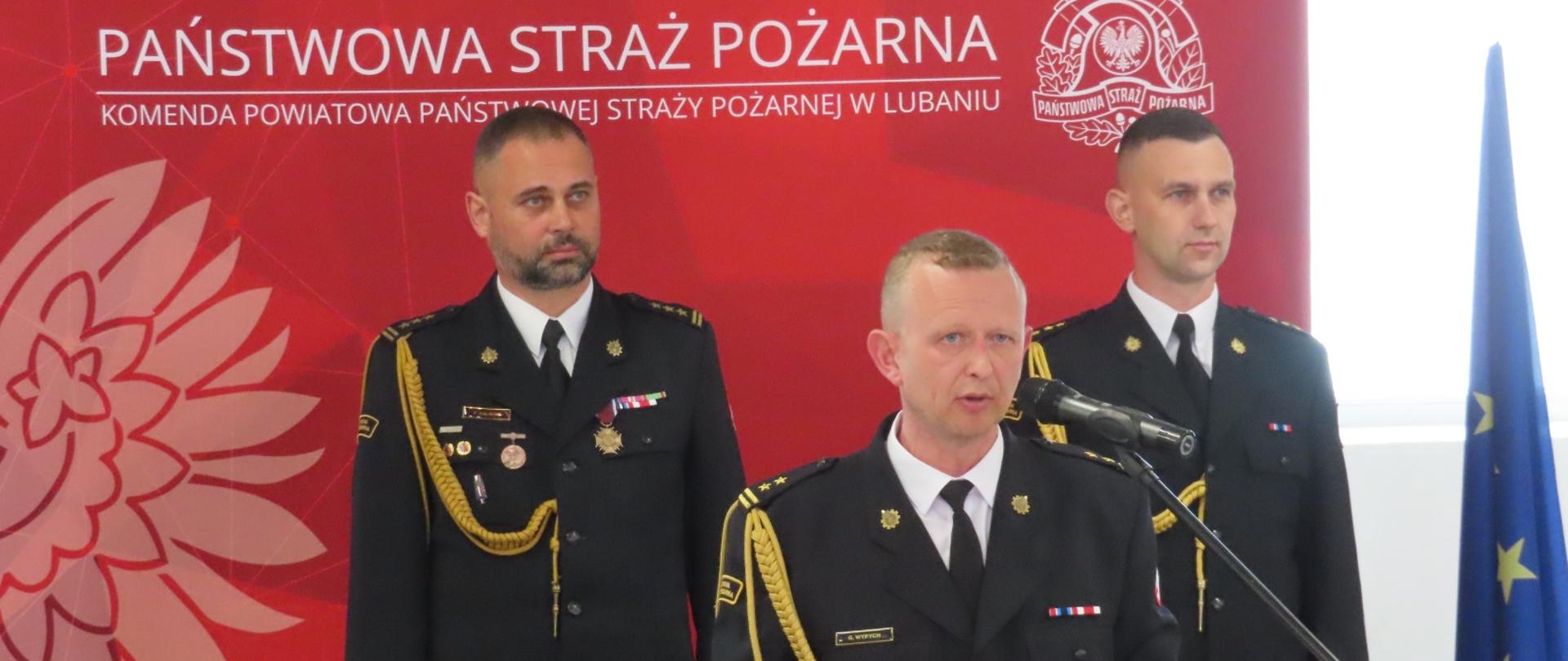Dzień Strażaka w Komendzie Powiatowej PSP w Lubaniu
Komendant Powiatowy wygłasza przemówienie, za nim stoją zastępca DKW i zastępca KP na czerwonym tle