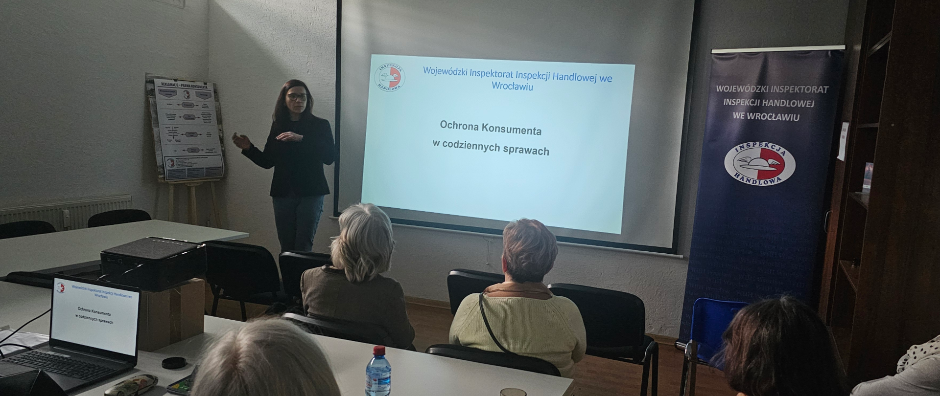 Sala szkoleniowa z rzutnikiem i ekranem, na którym wyświetlana jest prezentacja pt. " Ochrona konsumenta w codziennych sprawach." Przed ekranem stoi kobieta prowadząca wykład, gestykulując podczas mówienia. Na sali siedzi kilka osób, głównie kobiet, słuchających prezentacji. Obok ekranu znajduje się stojący banner Inspekcji Handlowej, a na stoliku stoi laptop, projektor i butelka wody. 