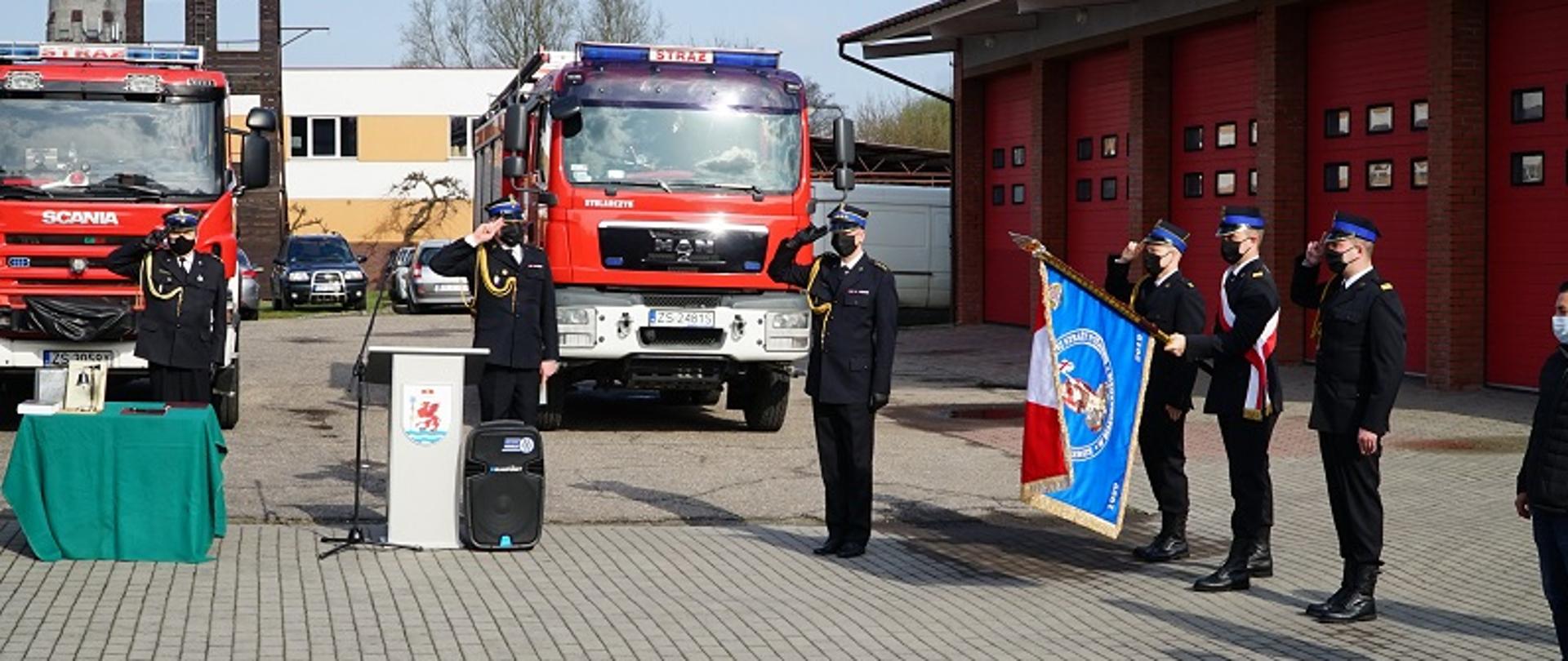 Zdjęcie przedstawia plac wewnętrzy KP PSP w Białogardzie. Na tle ustawionych przodem dwóch samochodów pożarniczych stoją od lewej: Komendant Powiatowy PSP w Białogardzie st. bryg. Jacek Szpuntowicz, dowódca JRG w Białogardzie bryg. Przemysław Zawadzki, z-ca dowódcy JRG w Białogardzie st. kpt. Mariusz Wenta. Z prawej strony trzyosobowy poczet sztandarowy. Strażacy w umundurowaniu wyjściowym stoją na baczność i salutują