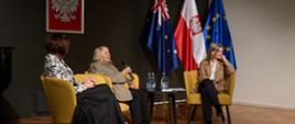 Trzy osoby na scenie, w tle flaga polska, australijska i unijna.