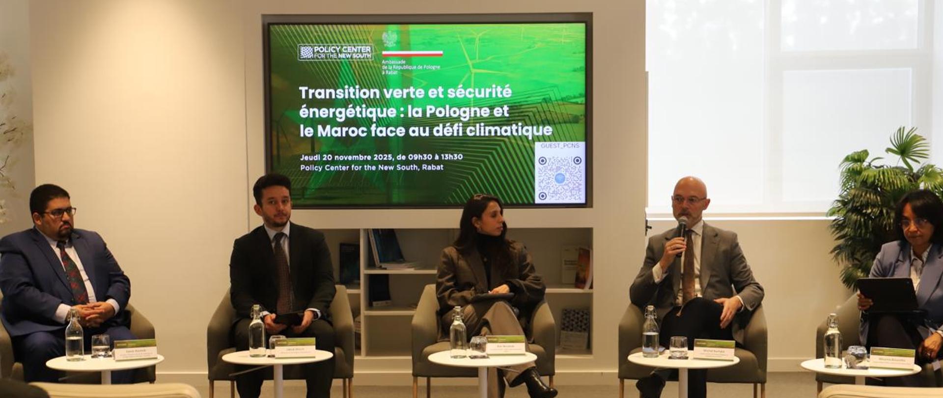 grupa osób siedzących w fotelach przy małych stolikach, jedna osoba trzyma mikrofon