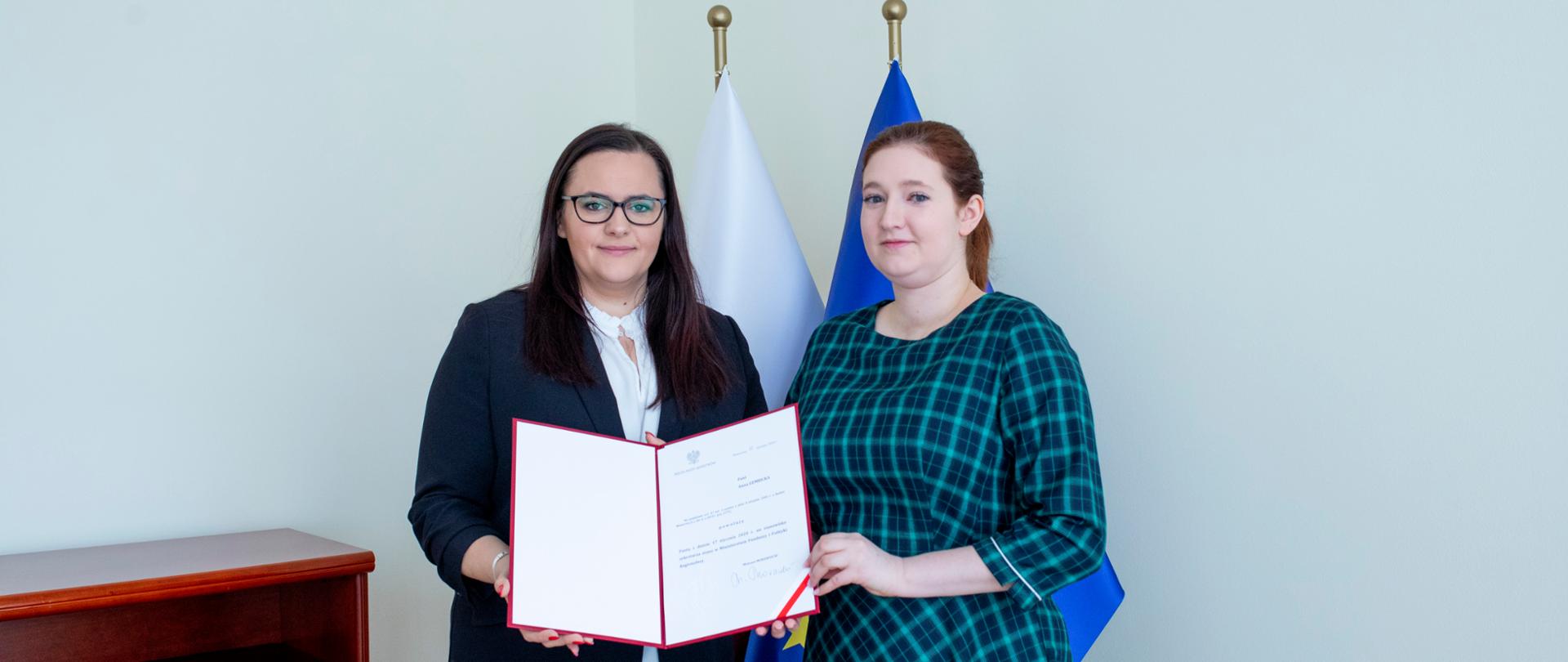 Minister Małgorzata Jarosińska-Jedynak i wiceminister Anna Gembicka trzymają nominację, za nimi flagi Polski i Unii Europejskiej