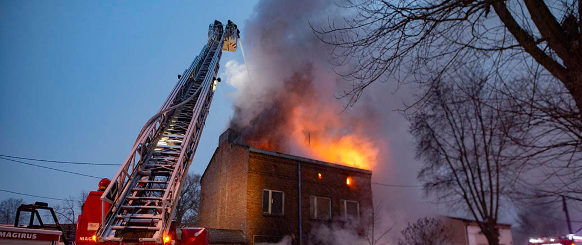 Na zdjęciu od lewej sprawiona drabina mechaniczna, w której koszu znajdują się dwaj strażacy podający z działka wodnopianowego prąd wody na palący się dach. Na prawo od drabiny grupa strażaków w aparatach układu oddechowego (OUO) przekazuje sobie informacje o pożarze. Przed grupą strażaków budynek z czerwonej cegły. Z bramy wjazdowej wydobywają się kłęby białego dymu. Po prawej stronie na skraju kadru pojazd gaśniczy z jednostki ratowniczo-gaśniczej w Pabianicach. 