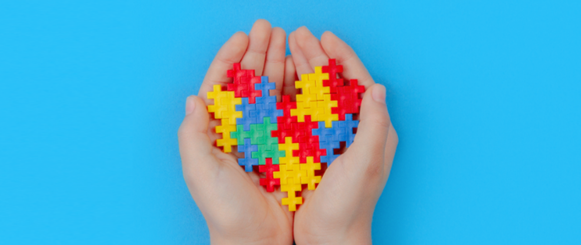 Na niebieskim tle złożone dłonie, w których ułożone jest serce z żółto-niebiesko-czerwono-zielonych puzzli