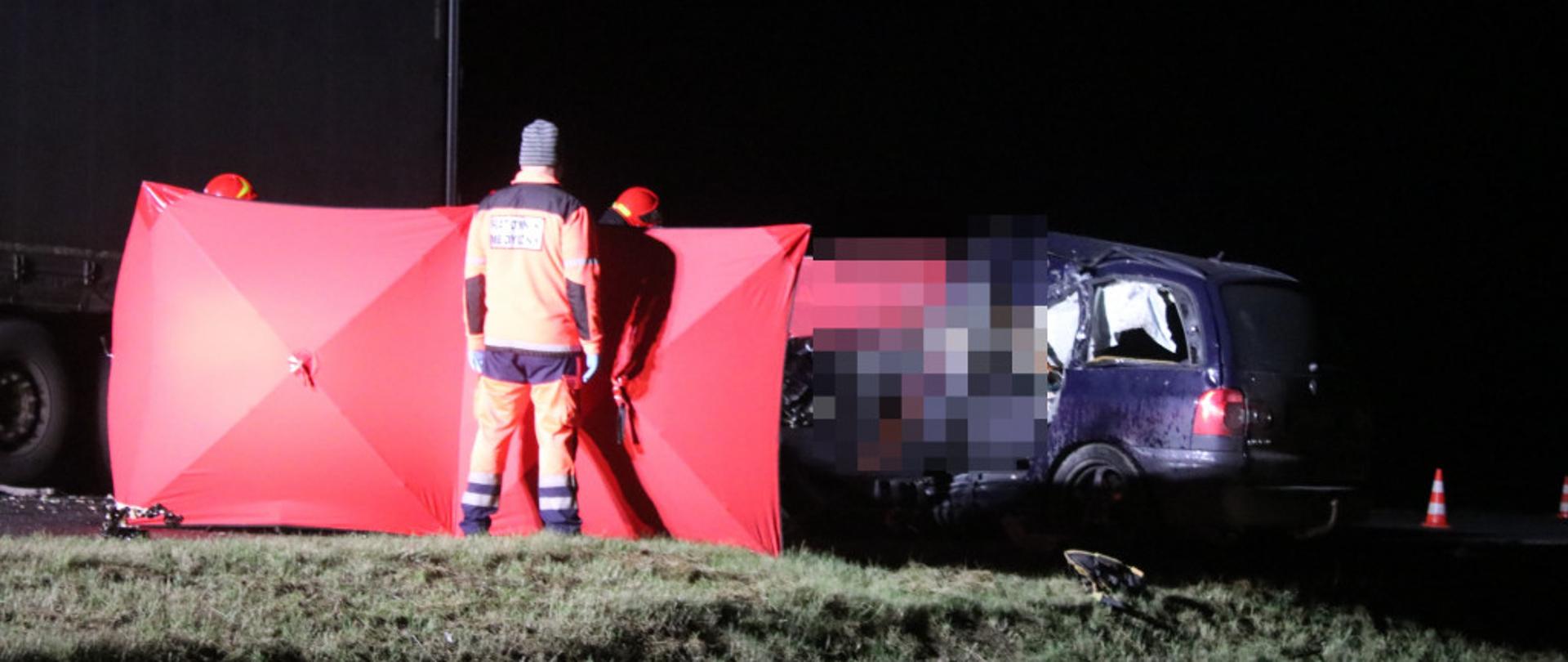 Zdjęcie przedstawia stojącego ratownika medycznego przed parawanem. Za nim znajduję się wrak pojazdu.