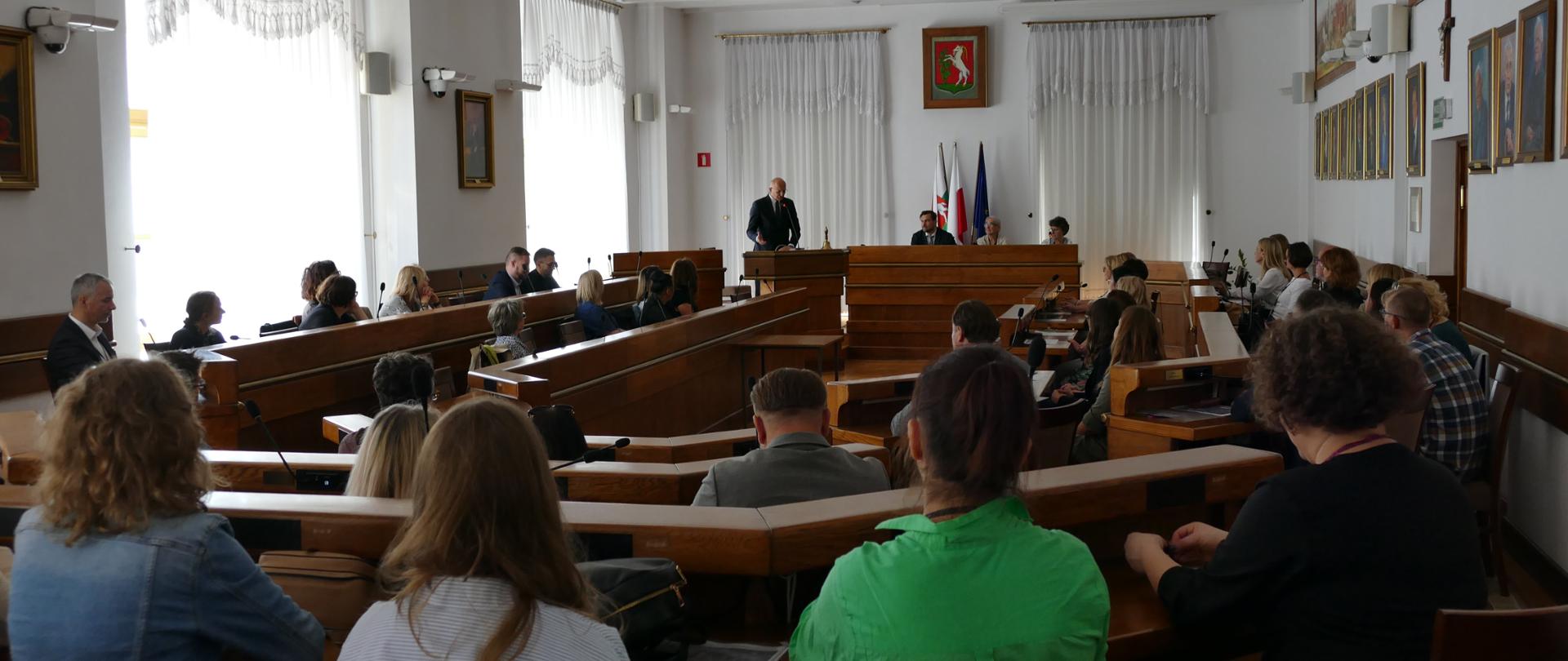 Biała sala, w której siedzą uczestnicy w ławach. W tle widać mężczyznę, który stoi za mównicą oraz stół prezydialny, za którym siedzą trzy osoby.