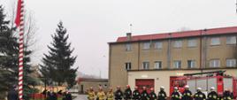 Strażacy Państwowej Straży Pożarnej podnoszą flagę Polski na maszt oddając honor, za nimi stoi samochód gaśniczy.