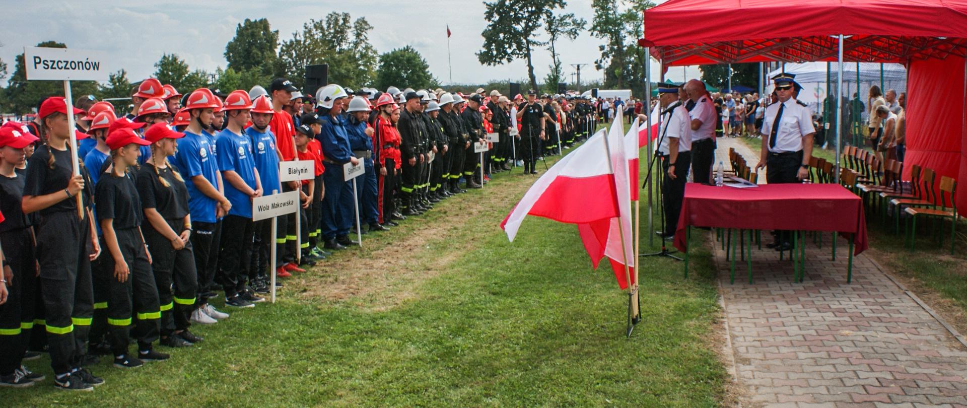 Na zdjęciu ustawione na boisku, w szeregu drużyny startujące w zawodach oraz zaproszeni goście. Tło stanowią biało-czerwone flagi i namiot koloru czerwonego.