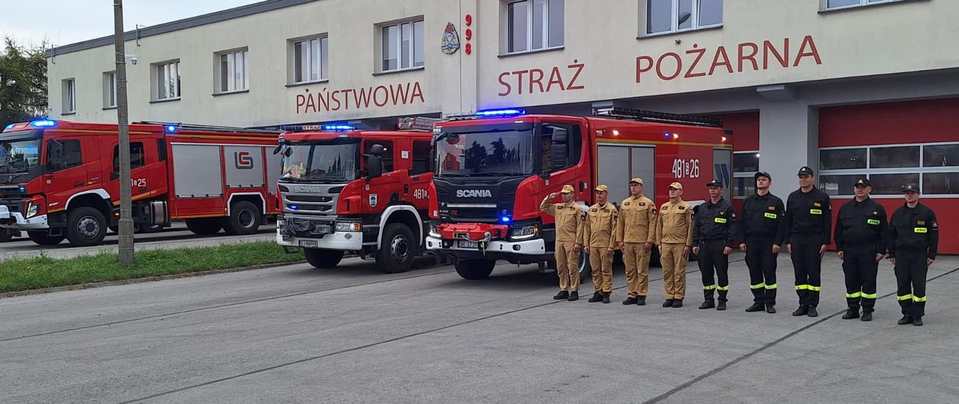 Strażacy w umundurowaniu koszarowym stoją w szyku przed budynkiem Komendy Miejskiej Państwowej Straży Pożarnej w Jaworznie. Obok nich zaparkowane są trzy wozy pożarnicze z włączonymi światłami błyskowymi.