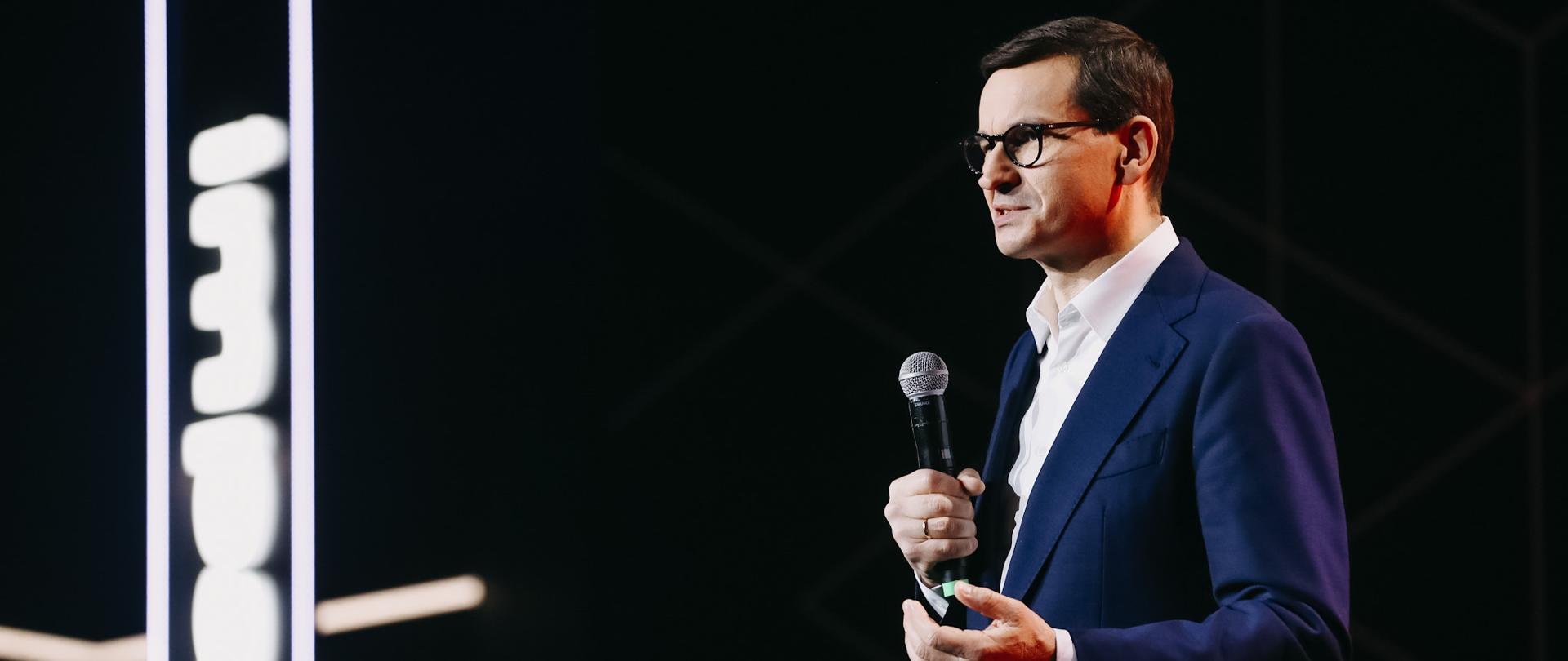 Premier stoi na scenie podczas przemówienia na tle napisu: impact '21.