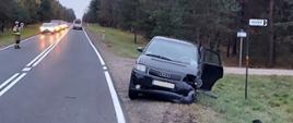 Uszkodzony samochód osobowy marki Audi stojący na poboczu drogi po zderzeniu z innym pojazdem. W aucie widoczne uszkodzenia zderzaka i lampy przedniej od strony kierowcy. W tle strażak kierujący ruchem.