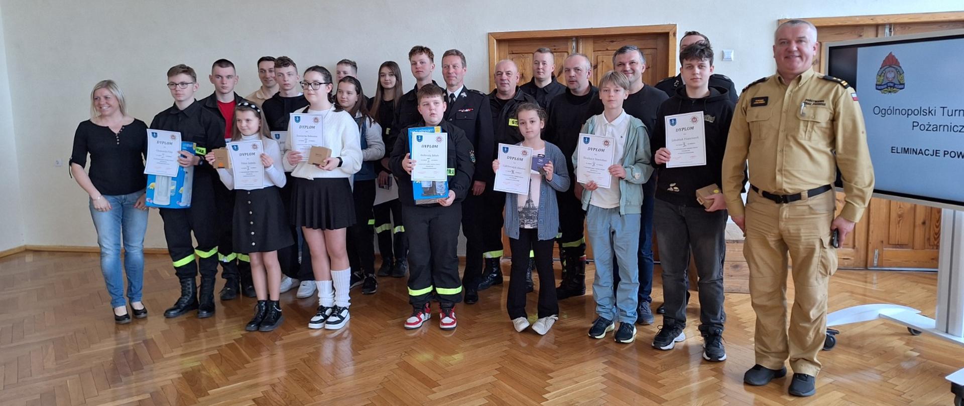 Uczestnicy wydarzenia z komisją egzaminacyjną i opiekunami. Zdjęcie wykonane w sali gimnastycznej strażnicy.