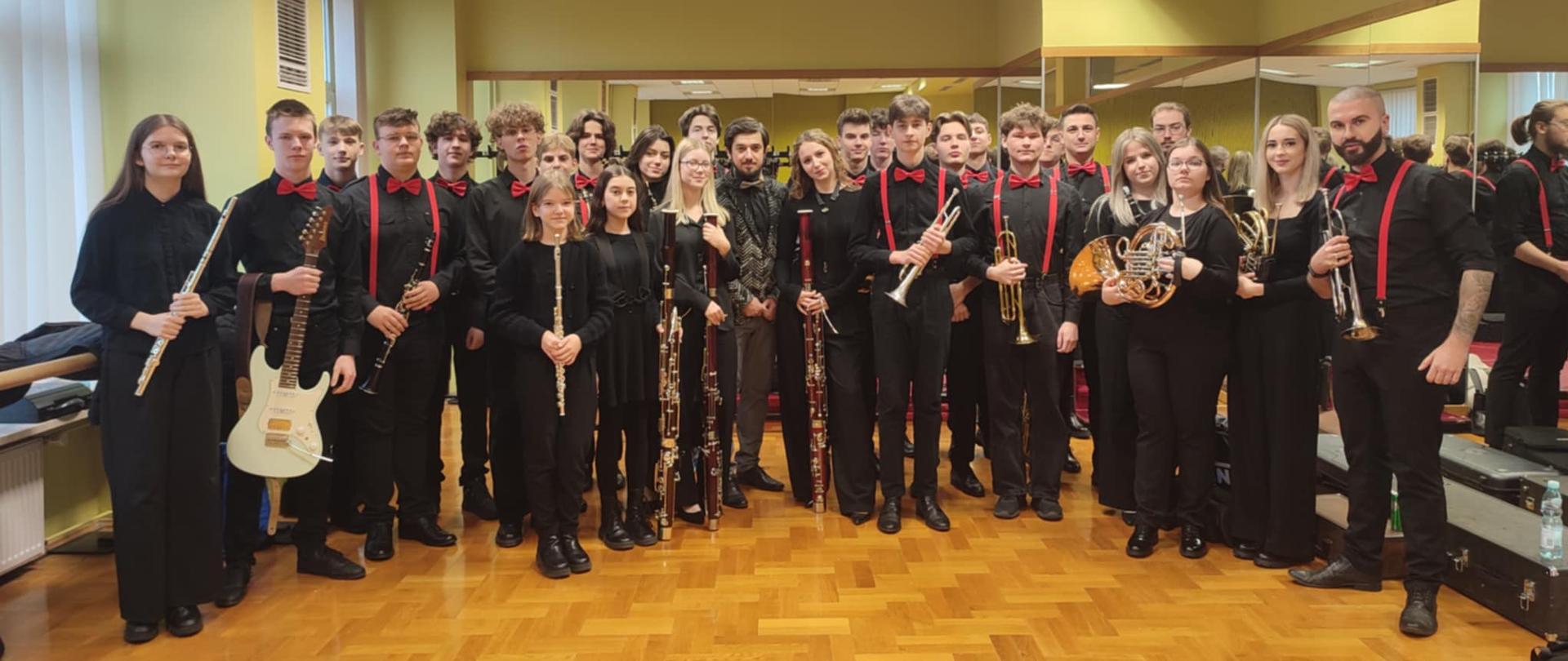 Grupa młodych muzyków z Orkiestry Dętej PSM II stopnia w Mielcu, ubrana w eleganckie czarne stroje z czerwonymi akcentami, pozuje do wspólnego zdjęcia przed koncertem w Centrum Kultury i Sztuki w Połańcu. Wśród nich znajdują się instrumentaliści grający na flecie, gitarze elektrycznej, klarnetach, trąbkach i waltorniach.