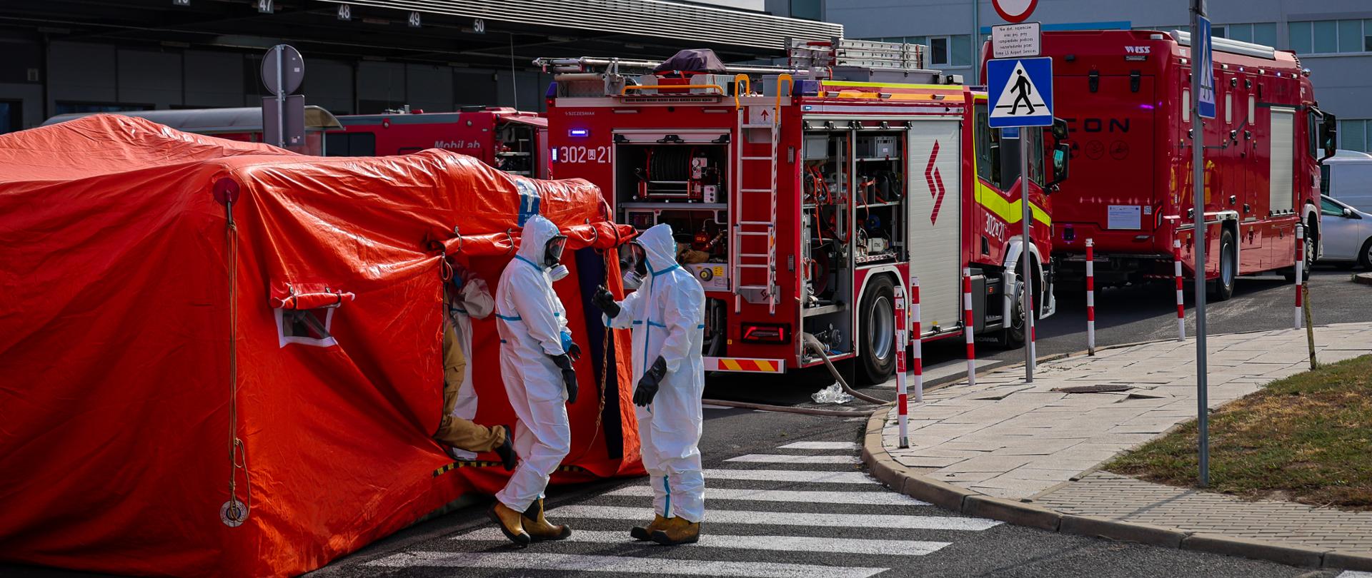 Widok na zewnątrz lotniska, gdzie trwa akcja ratunkowa. Centralnym punktem zdjęcia jest duży, czerwony namiot medyczny. Obok niego, na przejściu dla pieszych, stoi dwóch ratowników w białych kombinezonach ochronnych, rozmawiających ze sobą. W tle widać dwa czerwone wozy strażackie z napisem „STRAŻ POŻARNA” oraz budynki lotniskowe. Na pierwszym planie po prawej stronie znajduje się znak drogowy z okrągłym, czerwonym obramowaniem, a obok niego, na niebieskim tle, znak z białą strzałką skierowaną w górę.