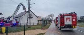 Na zdjęciu po lewej stronie widać budynek parterowy. Przy nim rozłożony podnośnik hydrauliczny, w którego koszu strażacy dogaszają sadzę w przewodzie kominowym. Po prawej stronie zaparkowane 3 pojazdy pożarnicze na poboczu drogi.