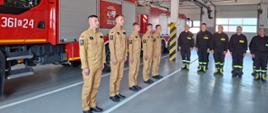 Na garażu obok pożarniczego samochodu bojowego w rzędzie stoją strażacy ubrani w piaskowe mundury. W tle inni strażacy w czarnych mundurach koszarowych