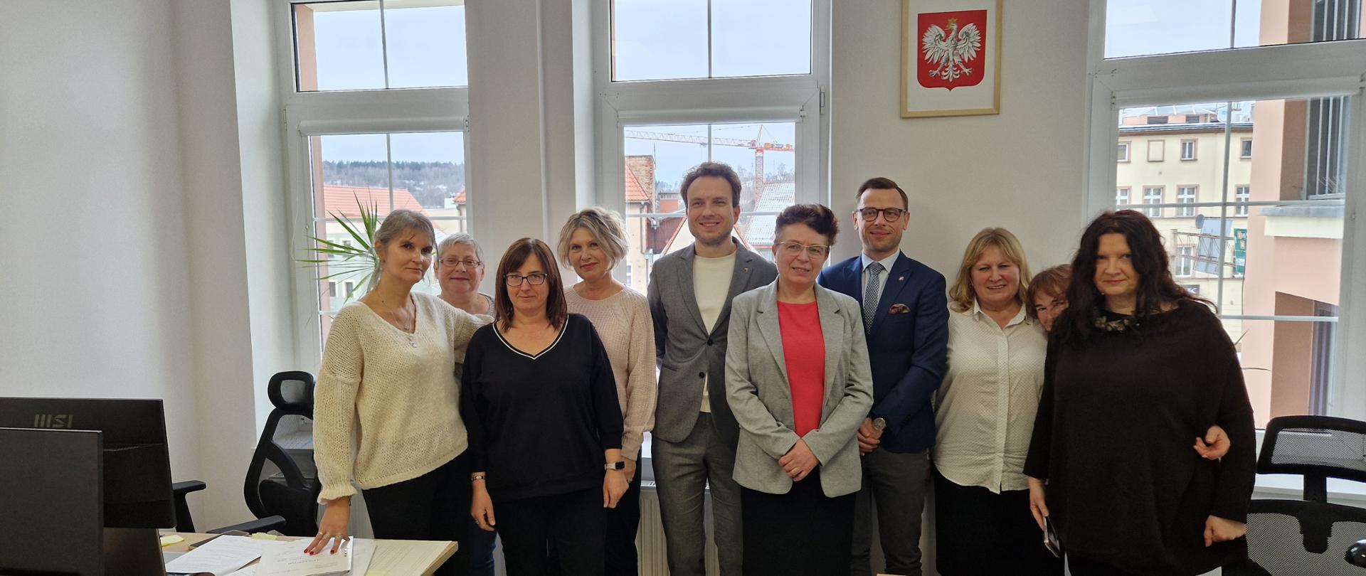Grupa dziewięciu dorosłych osób stoi w sali biurowej przy biurkach. Osoby ustawione są w dwóch rzędach i patrzą w stronę aparatu. Pomieszczenie jest jasne, z dużymi oknami w tle, na ścianie widoczny jest herb Polski. Na stołach leżą dokumenty i drobne przedmioty biurowe. 
