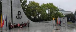 Na zdjęciu znajduje się szary pomnik z logo Polski Walczącej, przed którym stoją wiązanki z biało czerwonymi kwiatami. Z lewej strony widać trzy flagi: żółto czerwoną, biało czerwoną oraz granatową z żółtymi gwiazdami. Z prawej strony w tle znajduje się budynek Sejmu