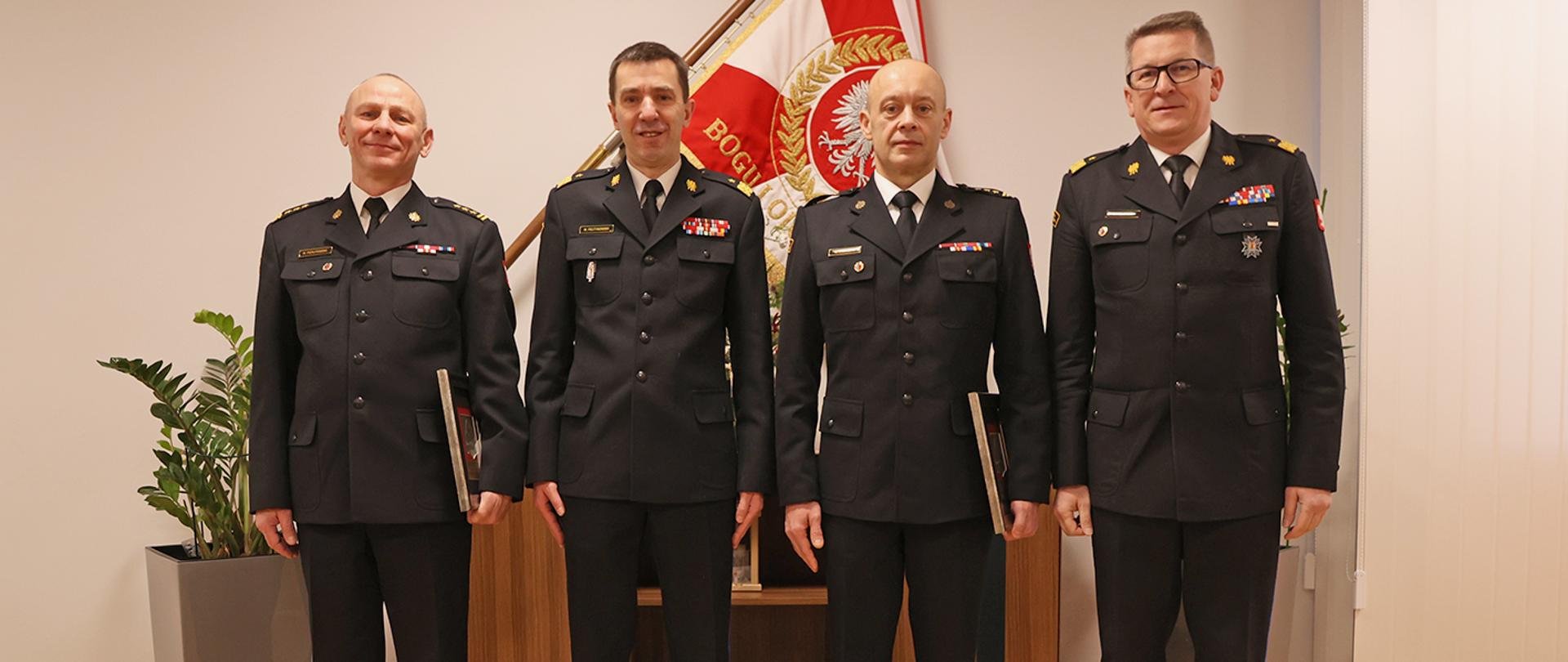 Zdjęcie pamiątkowe na tle sztandaru Komendy Głównej Państwowej Straży Pożarnej. Czterej mężczyźni w strażackich mundurach galowych stojący w postawie zadadniczej Dwaj mężczyźni w mundurach generalskich