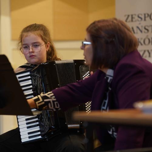 zdjęcie przedstawia uczennicę młodszej klasy grającą na akordeonie, z boku prowadząca warsztaty udziela wskazówek, w tle baner szkoły
