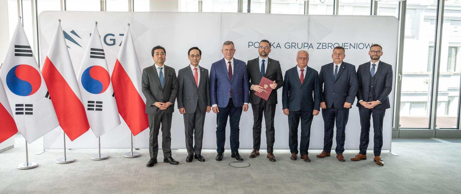 Family photo. Grupa osób na tle ścianki z logotypami PGZ. Z lewej strony flagi Polski i Korei Płd. 
