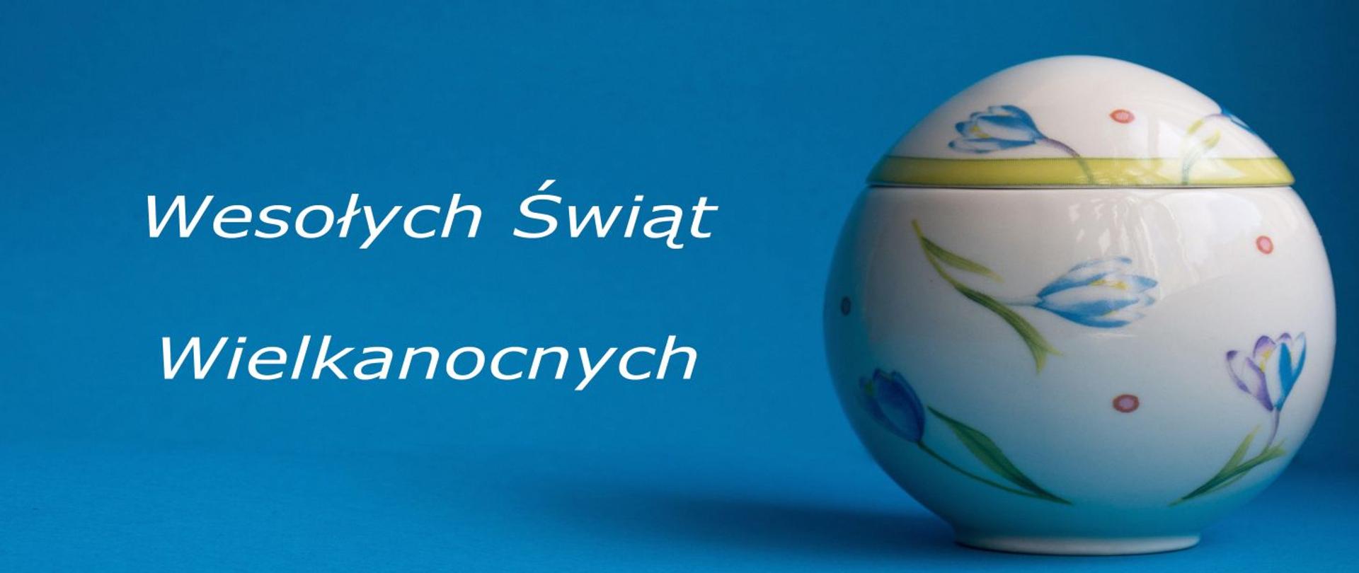 Grafika przedstawia biały napis Wesołych Świąt Wielkanocnych na niebieskim tle obok grafika szklanej pisanki