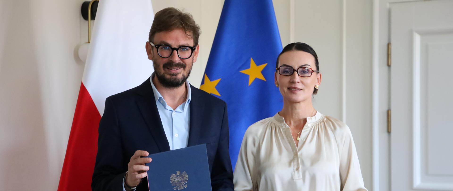 Dwie osoby stoją obok siebie w eleganckim wnętrzu, na tle flag Polski i Unii Europejskiej. Mężczyzna po lewej, ubrany w ciemny garnitur i jasną koszulę, trzyma granatową teczkę z godłem Polski. Kobieta po prawej ma na sobie jasną bluzkę i czarne spodnie. Oboje patrzą w stronę aparatu i lekko się uśmiechają.
