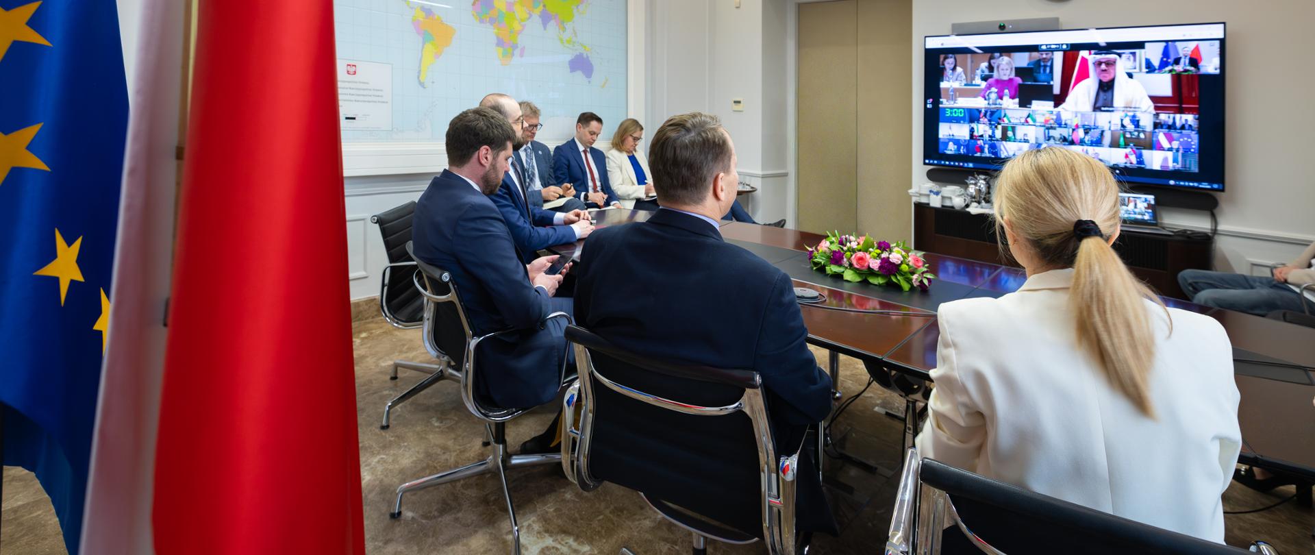 Siedem osób (dwie kobiety i pięciu mężczyzn) siedzi przy stole konferencyjnym podczas wideokonferencji. Na ekranie widocznych jest wielu uczestników spotkania online; w pomieszczeniu znajdują się flagi Polski i Unii Europejskiej oraz mapa świata na ścianie.