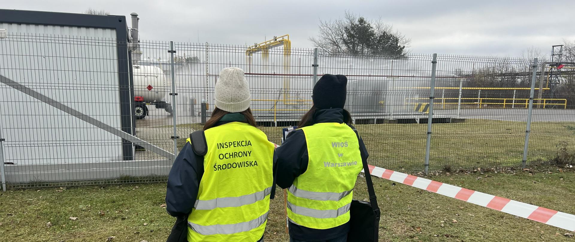 Dwie inspektorki Inspektoratu Ochrony Środowiska w Warszawie obserwują ćwiczenia przeciwpożarowe: w tle widać terminal gazu płynnego, na którym włączone są zraszacze.