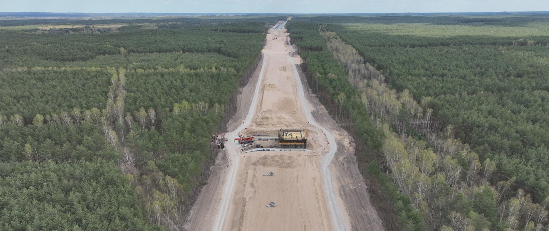 Widok z lotu ptaka na szeroki pas budowy drogi przebiegający przez gęsty las. W centrum kadru znajduje się prosty odcinek drogi w trakcie realizacji, pokryty jasnym piaskiem, z widocznymi śladami maszyn budowlanych oraz pojedynczymi betonowymi elementami ułożonymi na powierzchni. W połowie długości widoczny jest plac robót z konstrukcją mostową lub przepustem oraz sprzętem budowlanym, w tym dźwigiem. Po obu stronach przyszłej jezdni znajdują się tymczasowe drogi techniczne. Otaczający teren to rozległy, zielony las z przewagą drzew iglastych oraz pasami jaśniejszych drzew liściastych. Horyzont rozciąga się w oddali pod lekko zachmurzonym niebem. 