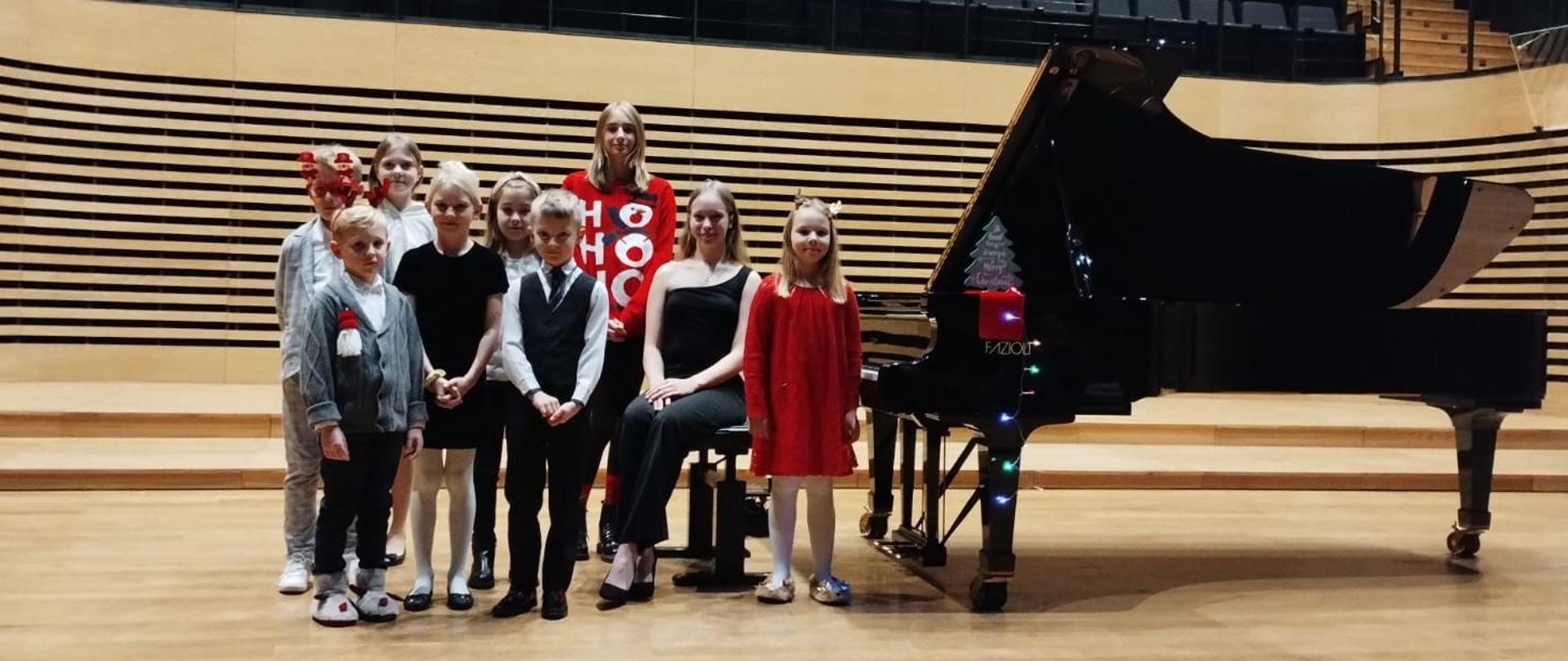 Na estradzie sali koncertowej nastolatka siedzi na krześle obok fortepianu stojącego po prawej stronie, wokół niej stoi pięć dziewczynek i trzech chłopców.